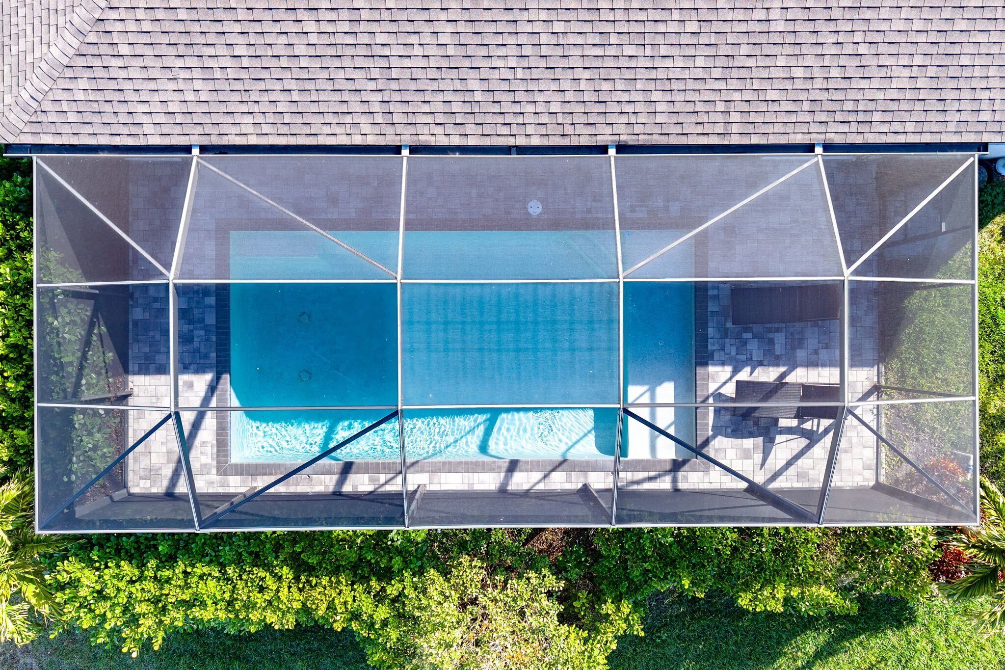 Ein rechteckiger Pool mit geometrischer Glaskonstruktion, umgeben von einer gefliesten Terrasse und üppiger Bepflanzung.