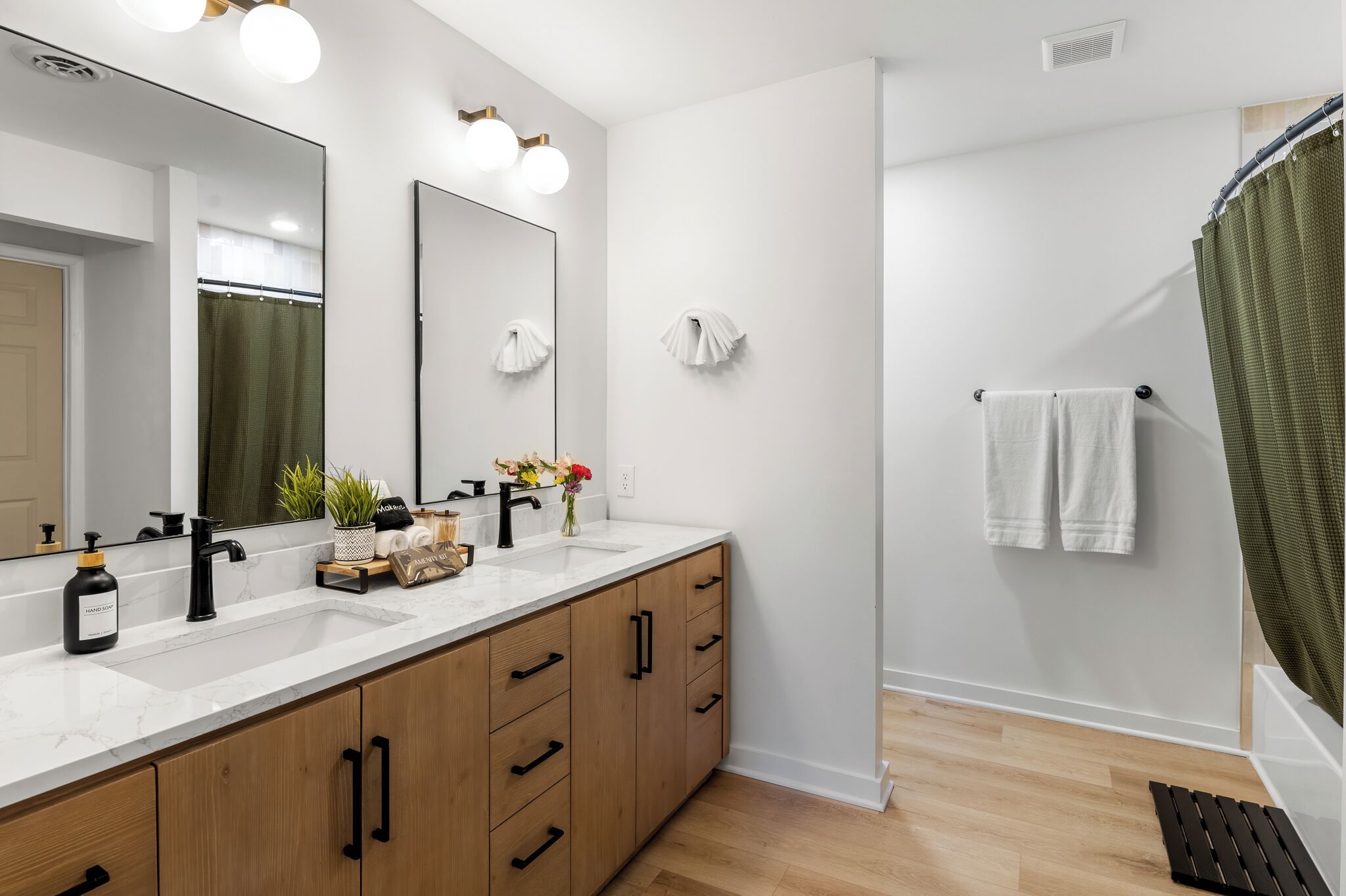 A modern bathroom with a white vanity, black faucets, and a large mirror. There are two towel racks on the wall and a shower curtain.