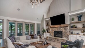 A spacious living room with a stone fireplace, a flat-screen TV mounted above it, and a comfortable seating area with sofas and armchairs. - Private Pool+Game Room+Private Dock on Lake Oconee (Eatonton)