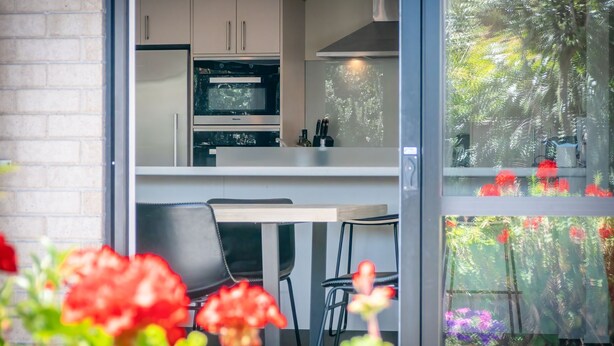 A modern kitchen with a built-in oven and microwave, a dining area with a table and chairs, and a view of a garden through a glass door. - A spacious family home in the centre of Mapua, close to everything but at the end of a quiet road (Mapua)