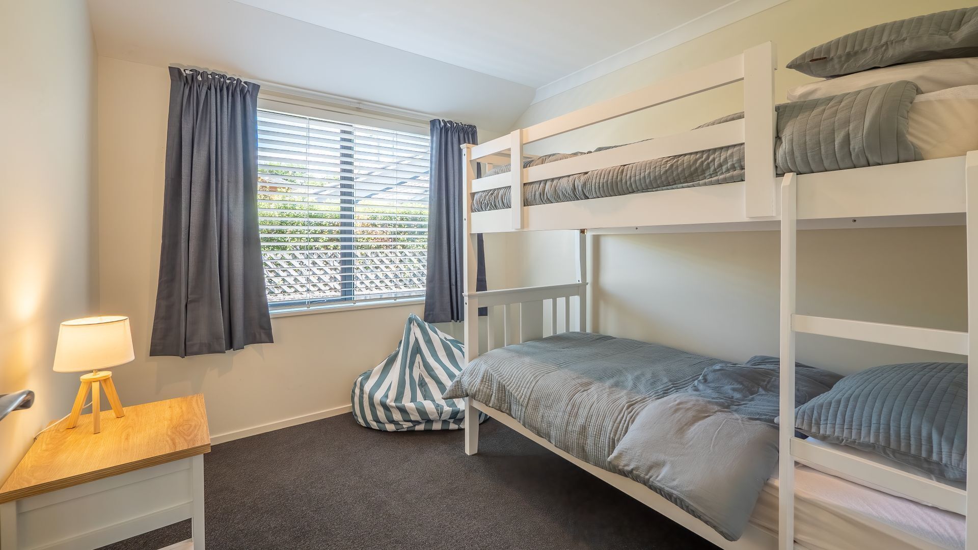 Une chambre avec un lit superposé, une fenêtre, des rideaux, un bureau avec une lampe et un coussin à rayures.