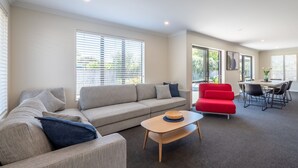 A modern living room with a grey sofa, a red chair, a wooden coffee table, and a dining area with a table and chairs. - A spacious family home in the centre of Mapua, close to everything but at the end of a quiet road (Mapua)