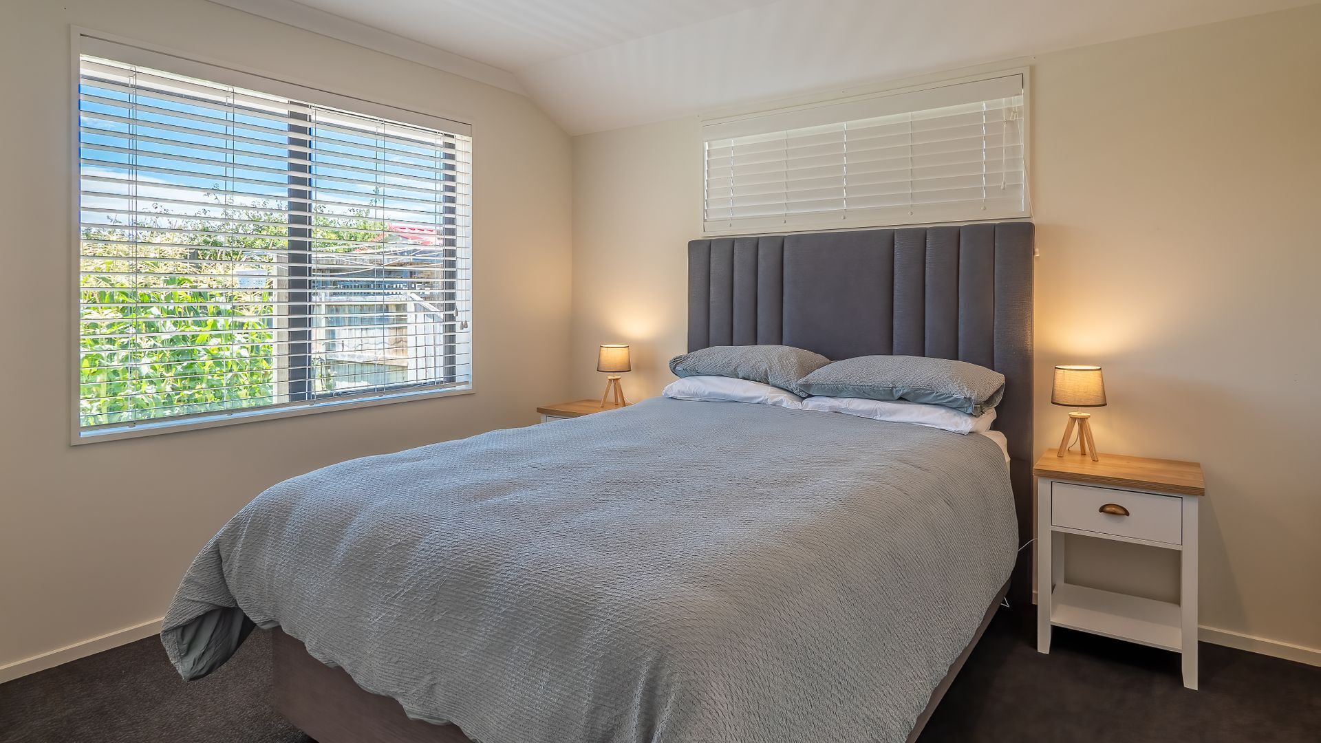 Quarto com uma cama grande, duas mesas de cabeceira com luminárias, uma janela com persianas e vista para áreas verdes.