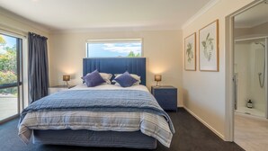 A bedroom with a large bed, a window with curtains, a nightstand with a lamp, a bathroom with a shower, and framed artwork on the wall. - A spacious family home in the centre of Mapua, close to everything but at the end of a quiet road (Mapua)
