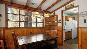 A wooden dining table with chairs, a kitchen area with a refrigerator, and a window with blinds.