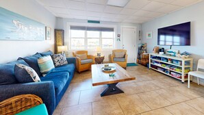 A living room with a blue sofa, two armchairs, a coffee table, and a bookshelf filled with books and games.