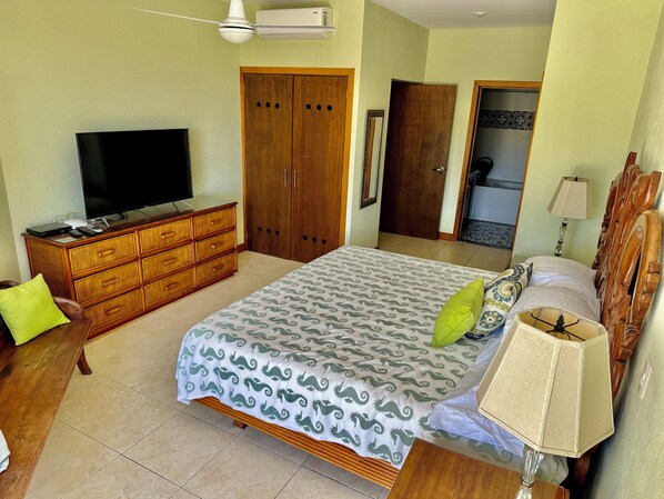 Un dormitorio con cama, cómoda, televisor, armario y baño a la vista a través de una puerta abierta.