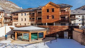 A ski resort with wooden buildings, a glass-roofed pool, and a snowy landscape. - Appt Cosy | 500m des Pistes | Piscine & sauna (Abriès)