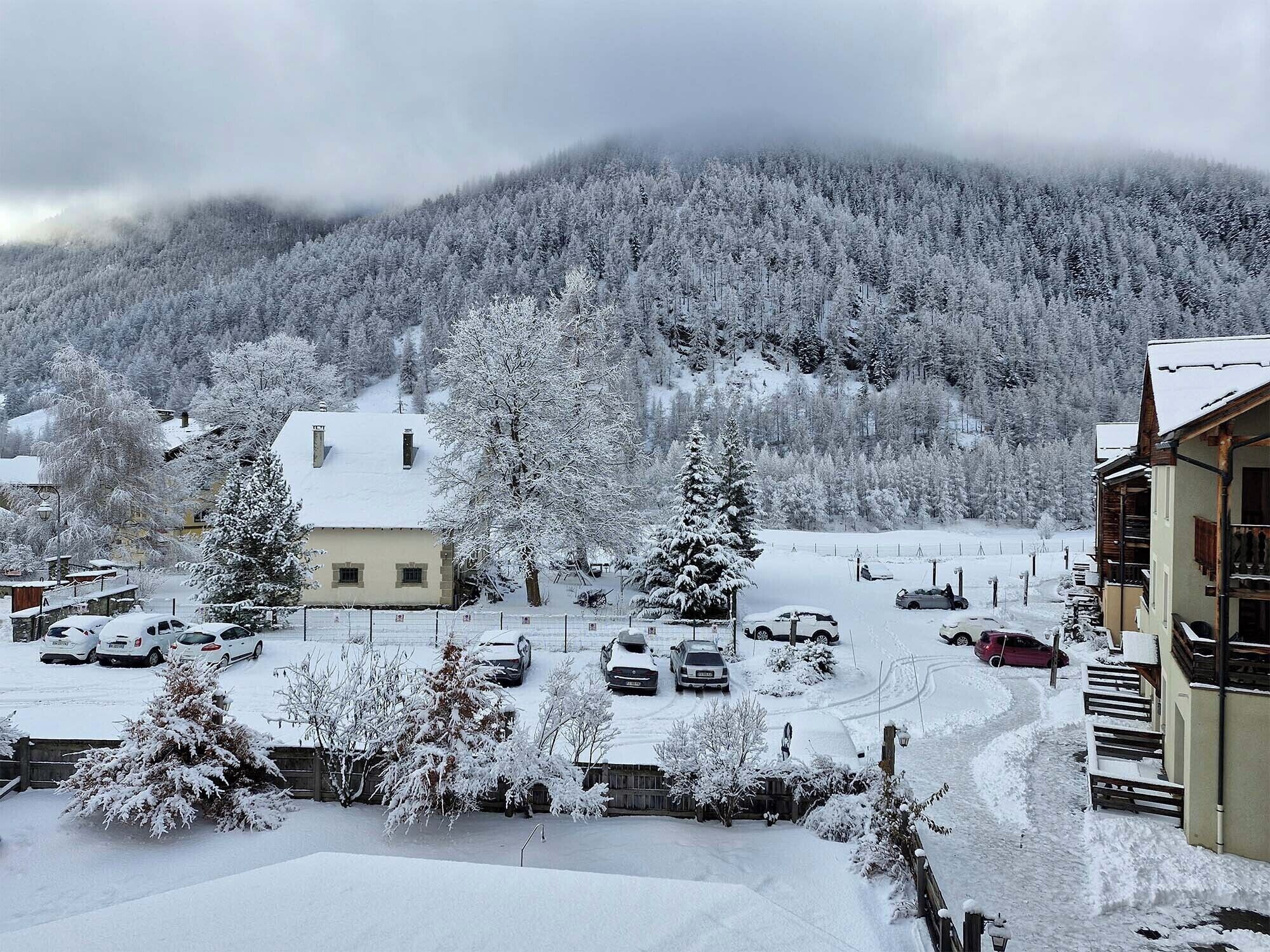 Ein verschneites Wohngebiet mit Häusern, parkenden Autos und einem bewaldeten Berg im Hintergrund.
