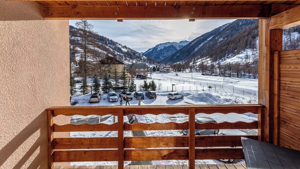 A wooden deck with a view of a snowy mountain landscape, including a road and parked cars. - Escapade alpine | Piscine, sauna & vue montagne (Abriès)