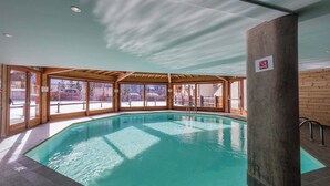An indoor swimming pool with clear blue water, wooden walls, and large windows. - Escapade alpine | Piscine, sauna & vue montagne (Abriès)