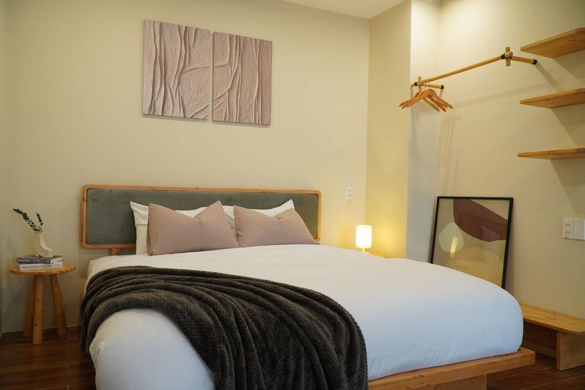 A neatly made bed with a dark throw blanket, a bedside table with a book and a vase, and a wall-mounted wooden rack with clothes.
