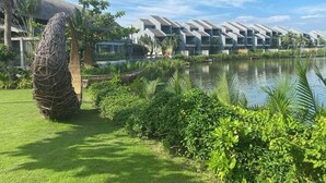 Ett modernt hotell med en unik arkitektonisk design, bestående av en serie sammanlänkade byggnader med en reflekterande yta, beläget vid en lugn sjö omgiven av frodig grönska och en byggnad med stråtak.