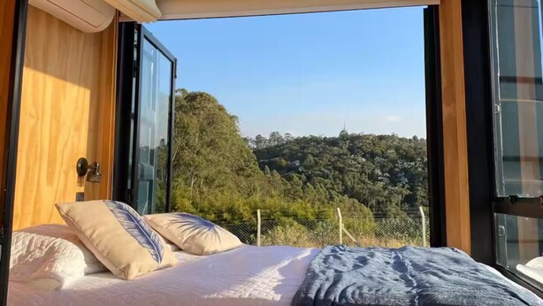 A bedroom with a large skylight, a bed with pillows, and a view of a forested area. - Cozy cabin with the best sunset! (Morungaba)