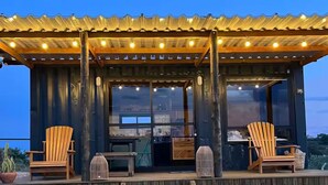 A wooden deck with two chairs and a table, a covered area with lights, and a view of the ocean. - Cozy cabin with the best sunset! (Morungaba)