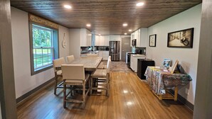 Une cuisine spacieuse avec un plancher en bois, un coin repas avec une grande table et des chaises, et une vue sur l’extérieur par une fenêtre.