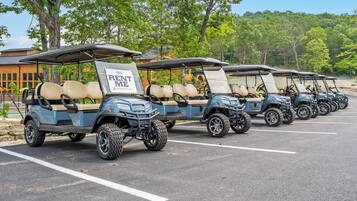 A row of golf carts parked in a lot with 'RENT ME' signs, surrounded by trees and a building in the background.