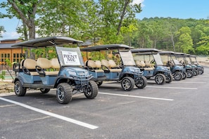 Una fila de carritos de golf estacionados en un estacionamiento con letreros que dicen 'ALQUÍLAME', rodeados de árboles y un edificio al fondo.