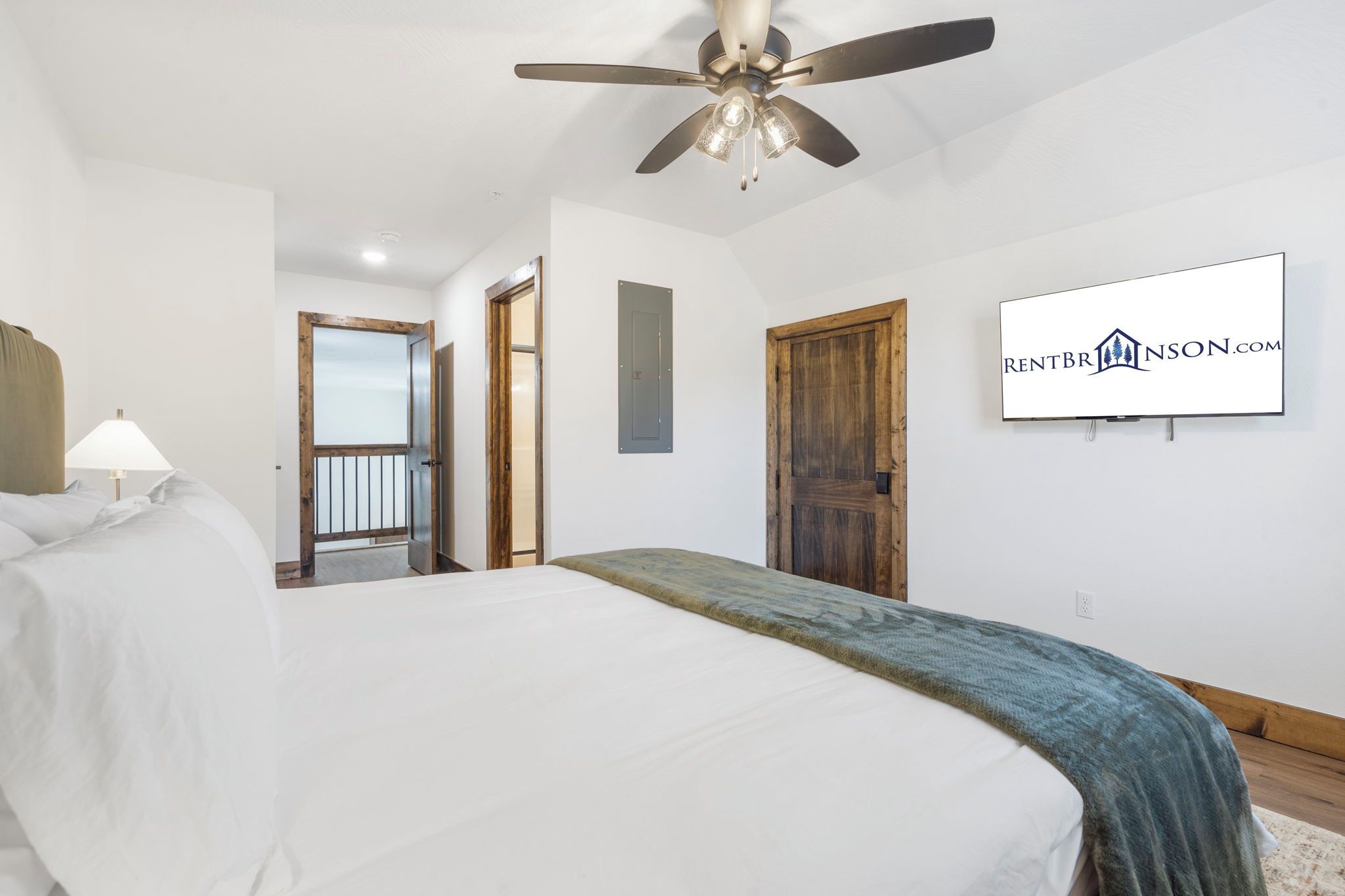 A bedroom with a bed, a ceiling fan, a wall-mounted TV, and a door leading to another room.