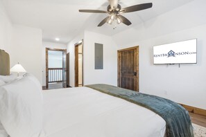 A bedroom with a bed, a ceiling fan, a wall-mounted TV, and a door leading to another room.