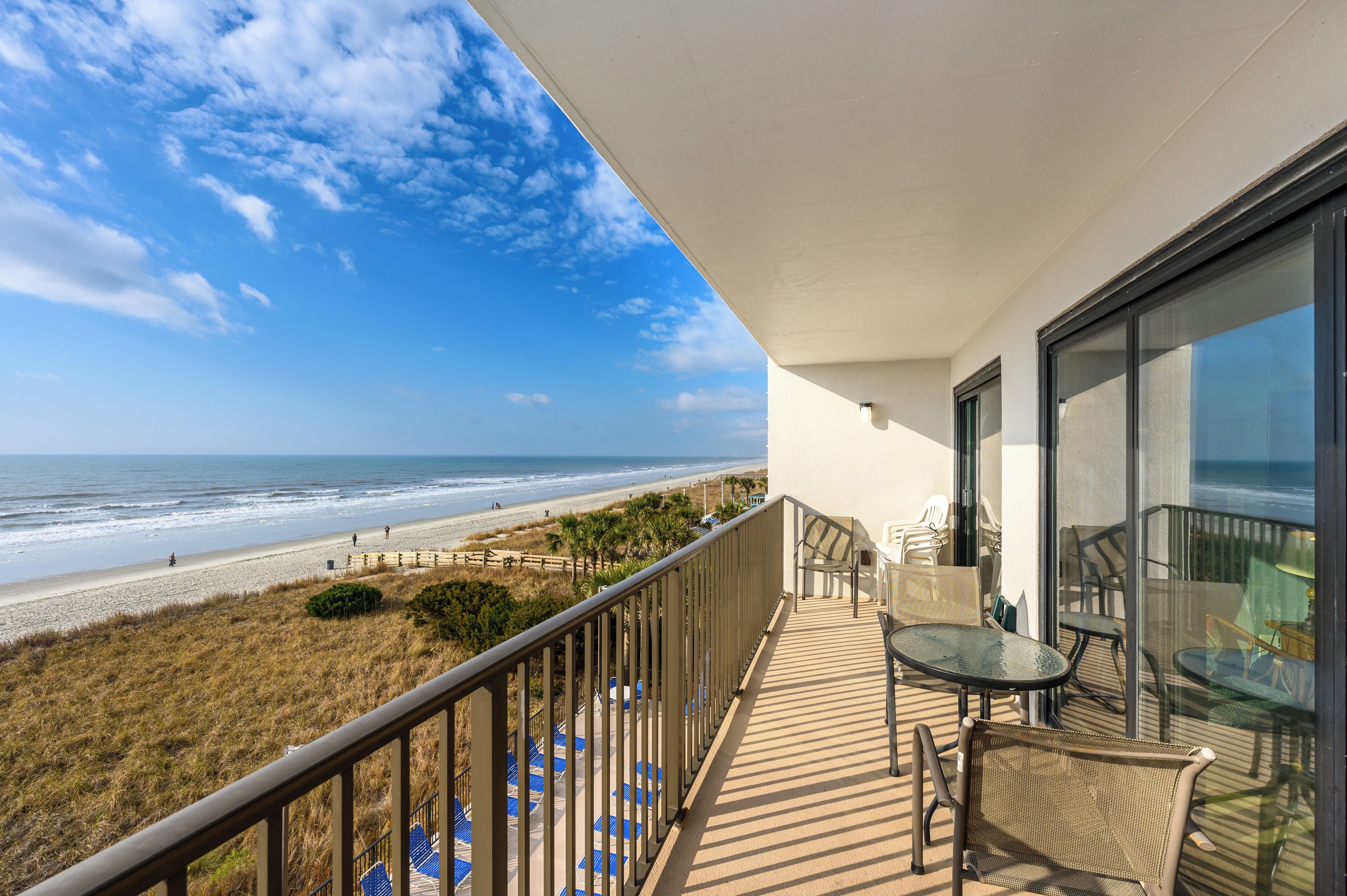 Un balcón con vista a la playa, mobiliario exterior y una puerta de vidrio que da a un patio.