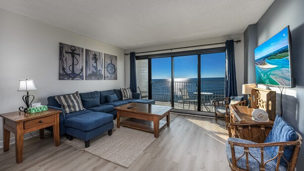 A modern living room with a blue sofa, wooden coffee table, and a large TV screen displaying a scenic view. - Oceanfront Pool-3BD Family Friendly Condo-Cherry Grove (North Myrtle Beach)