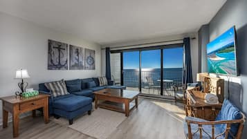 Una sala moderna con un sofá azul, una mesa de centro de madera y una gran pantalla de televisión mostrando un paisaje pintoresco.