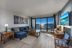 Een moderne woonkamer met een blauwe bank, een houten salontafel en een groot televisiescherm dat een mooi landschap toont.