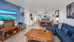 A spacious living room with a large flat-screen TV, a wooden coffee table, a blue sofa, and a dining area with a wooden table and chairs. - Oceanfront Pool-3BD Family Friendly Condo-Cherry Grove (North Myrtle Beach)