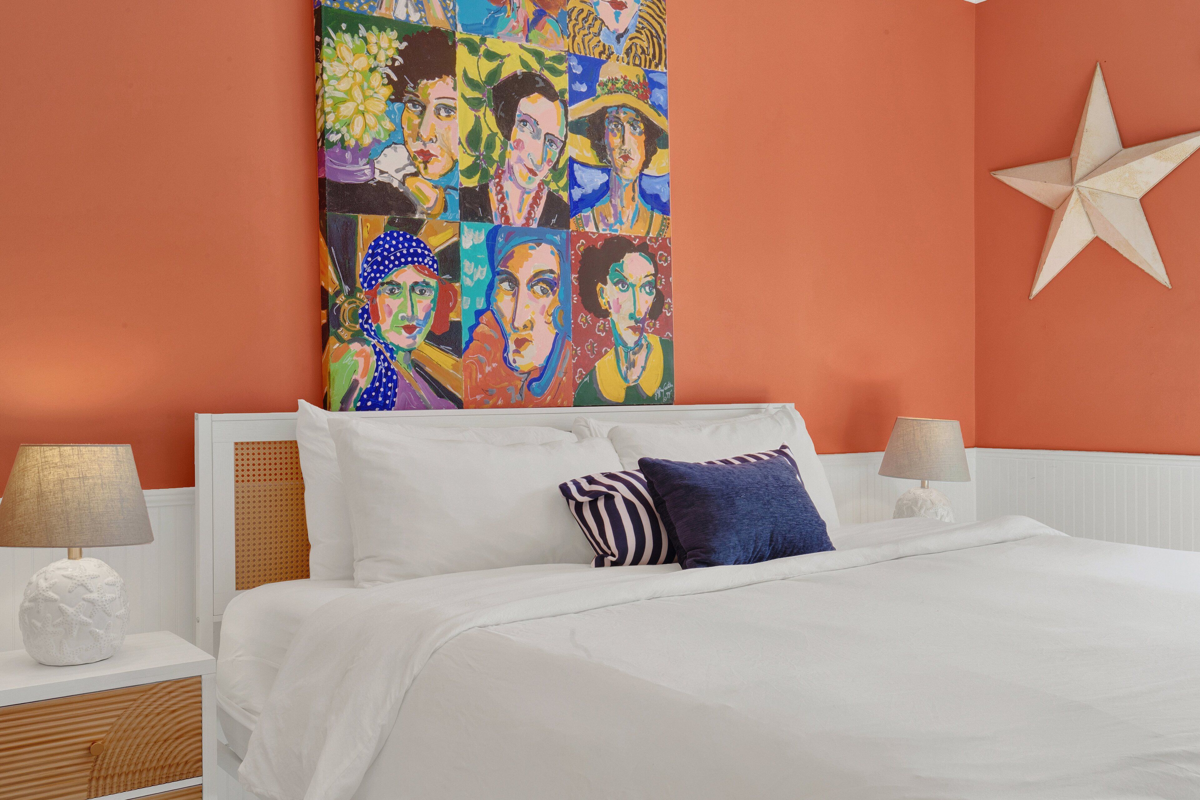 Een slaapkamer met een wit bed, gestreepte kussens, een sterren decoratie aan de muur en een kleurrijk kunstwerk boven het bed.