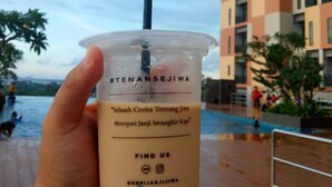 A hand holding a plastic cup with a straw, labeled with a hashtag and a slogan, in front of a building with a pool and people in the background.