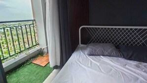 A bedroom with a bed, a window with curtains, a small balcony with a railing, and a wall-mounted air conditioning unit.