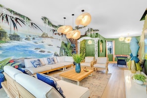 A living room with a beach mural, a sofa set, a coffee table, and a dining area with a table and chairs. - Themed Glow Villa • 4K Cinema • Heated Pool • Lake • Sleeps 12 (Miami)