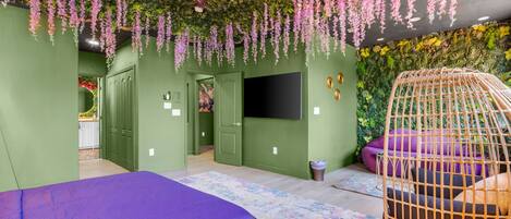 A room with a bed, a television, a hanging plant, and a wicker chair.