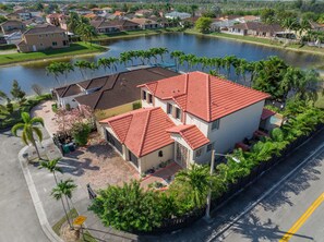 A residential area with a house featuring a red-tiled roof, surrounded by a small pond and palm trees. - Themed Glow Villa • 4K Cinema • Heated Pool • Lake • Sleeps 12 (Miami)