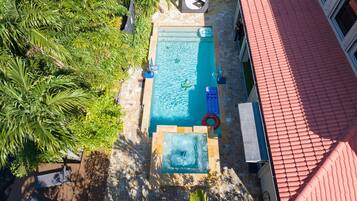 An aerial view of a swimming pool with a tiled deck, surrounded by lush greenery and a red-roofed building.