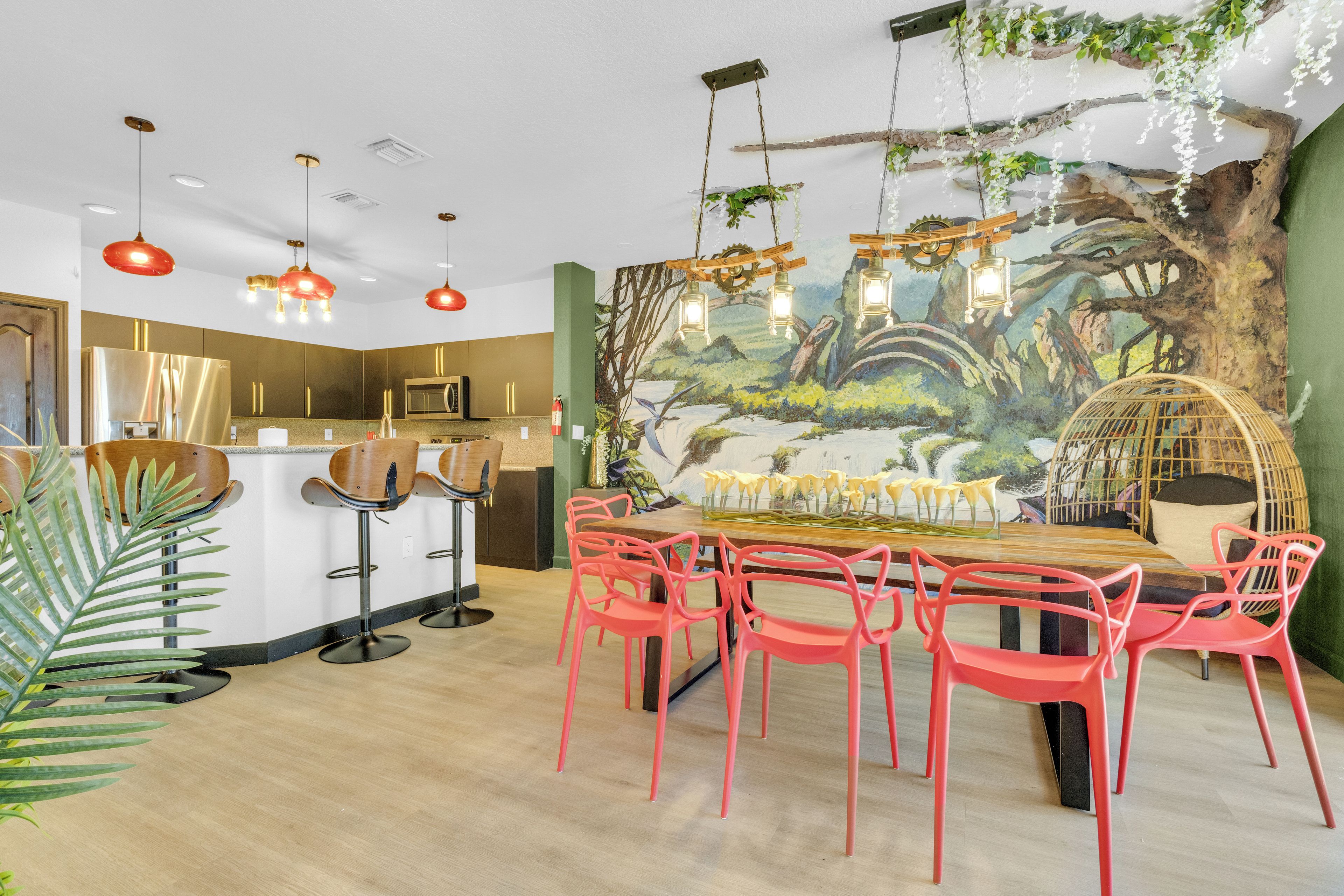 Uma área de refeições com mesa de madeira e cadeiras vermelhas, um mural com árvore e flores, e luminárias pendentes.