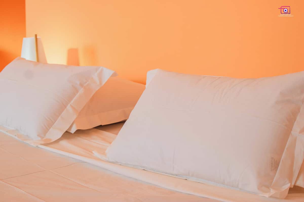 Uma cama com roupa de cama e almofadas brancas, uma mesa de cabeceira e um abajur fixado na parede.