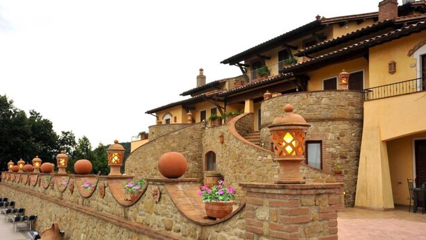 Un mur de pierre agrémenté de pots en terre cuite et de lanternes, un bâtiment à la façade jaune et doté d’un balcon, et un espace pavé avec des chaises.