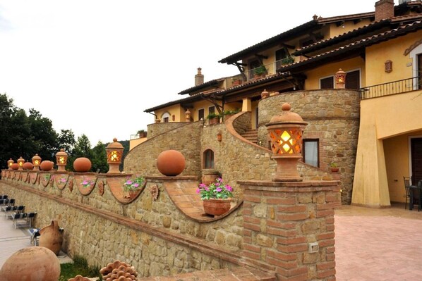 A stone wall with terracotta pots and lanterns, a building with a yellow facade and a balcony, and a paved area with chairs.