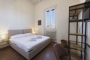 Quarto com cama, luminárias de cabeceira, radiador, janela e uma cadeira.
