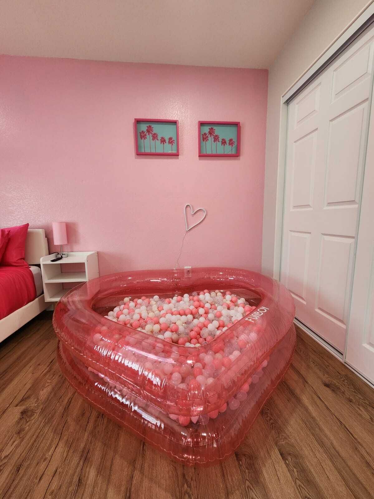 Une chambre décorée dans des tons roses, avec un lit recouvert d’une literie rouge, une table de chevet blanche et un mur orné de deux tableaux encadrés.