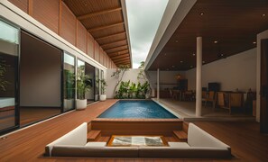 A modern indoor pool area with wooden flooring, a seating area with cushions, and potted plants. - Kinama Villa (Jimbaran)