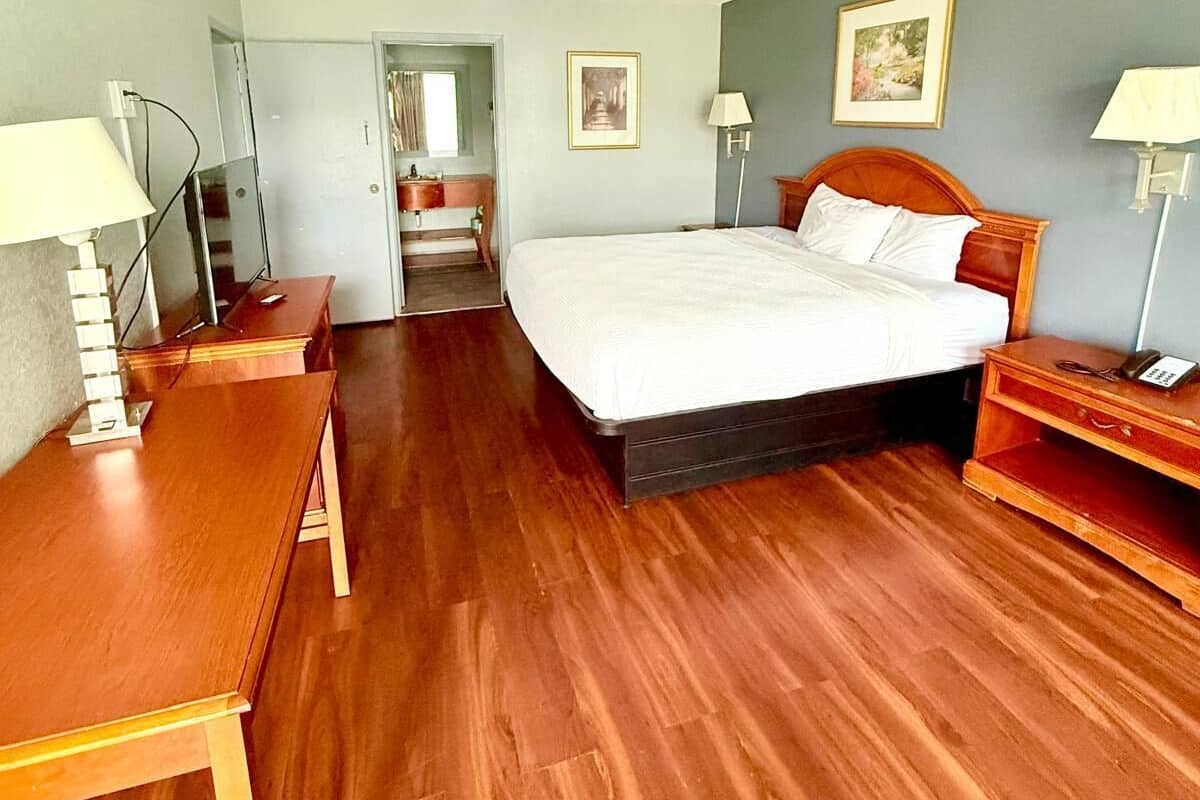 A hotel room with a wooden floor, a bed with white bedding, two bedside tables with lamps, a desk, and a chair.