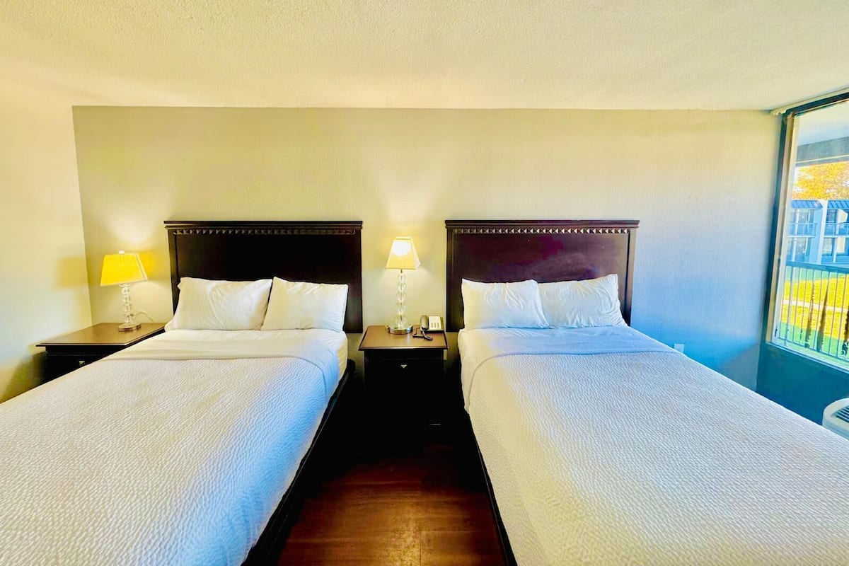 A hotel room with two beds, each with a wooden headboard, white bedding, and a nightstand with a lamp.