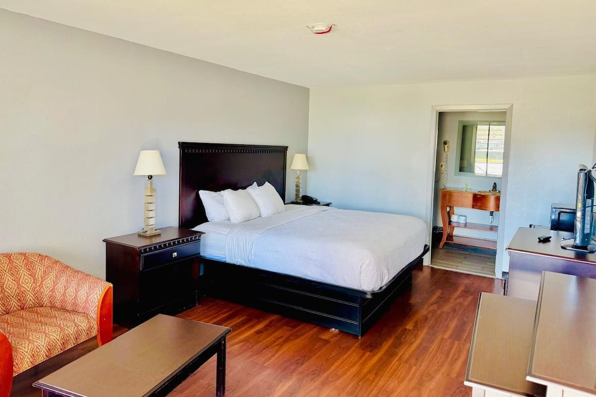 A hotel room with a large bed, two bedside tables with lamps, a wooden chair, a small table, and a desk with a computer.