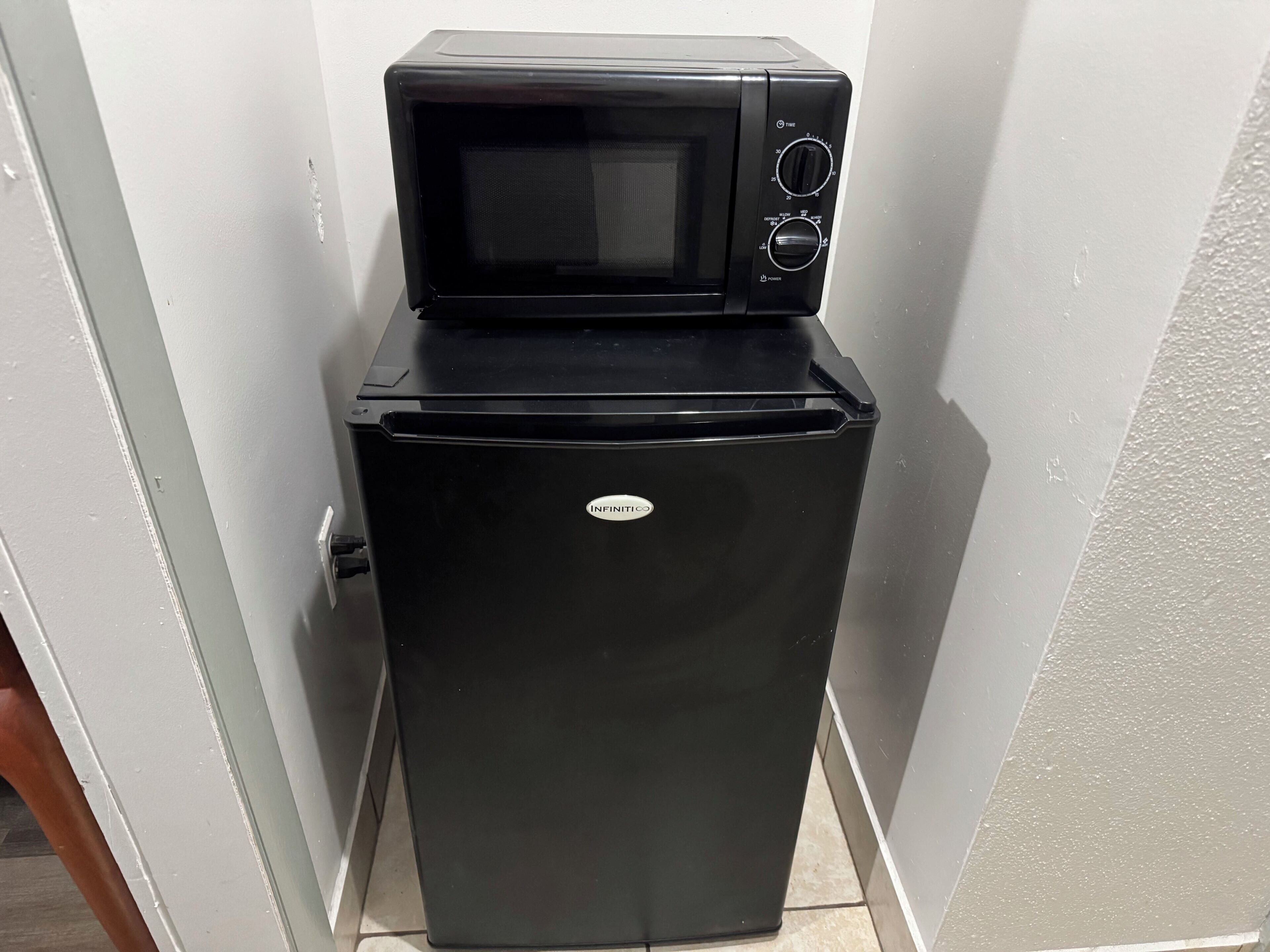 A black refrigerator with a microwave on top, placed in a corner of a room with white walls and a tiled floor.