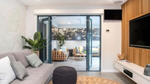 Una sala de estar moderna con un sofá, una mesa de centro y un televisor. Hay un ventanal corredero que da a un balcón con mobiliario exterior.