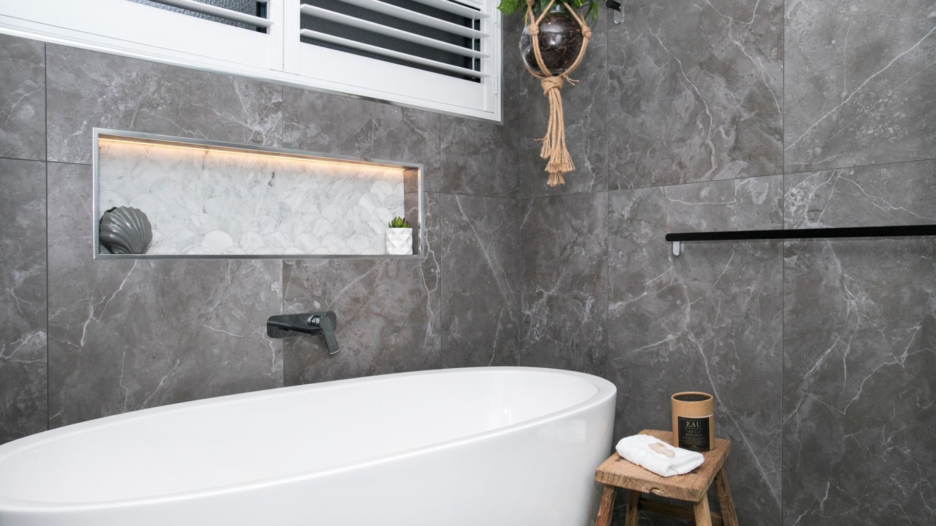 A modern bathroom with a freestanding bathtub, a small wooden stool with a towel, a potted plant, and a framed mirror.