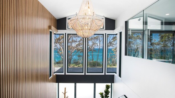 A modern interior with a large chandelier, a view of the sea through large windows, and a wooden slat wall. - 5 BDR - 250m to Beach - Outdoor Spa - Book 3 Nights, Pay for 2 until 24 September 2026 (Hyams Beach)